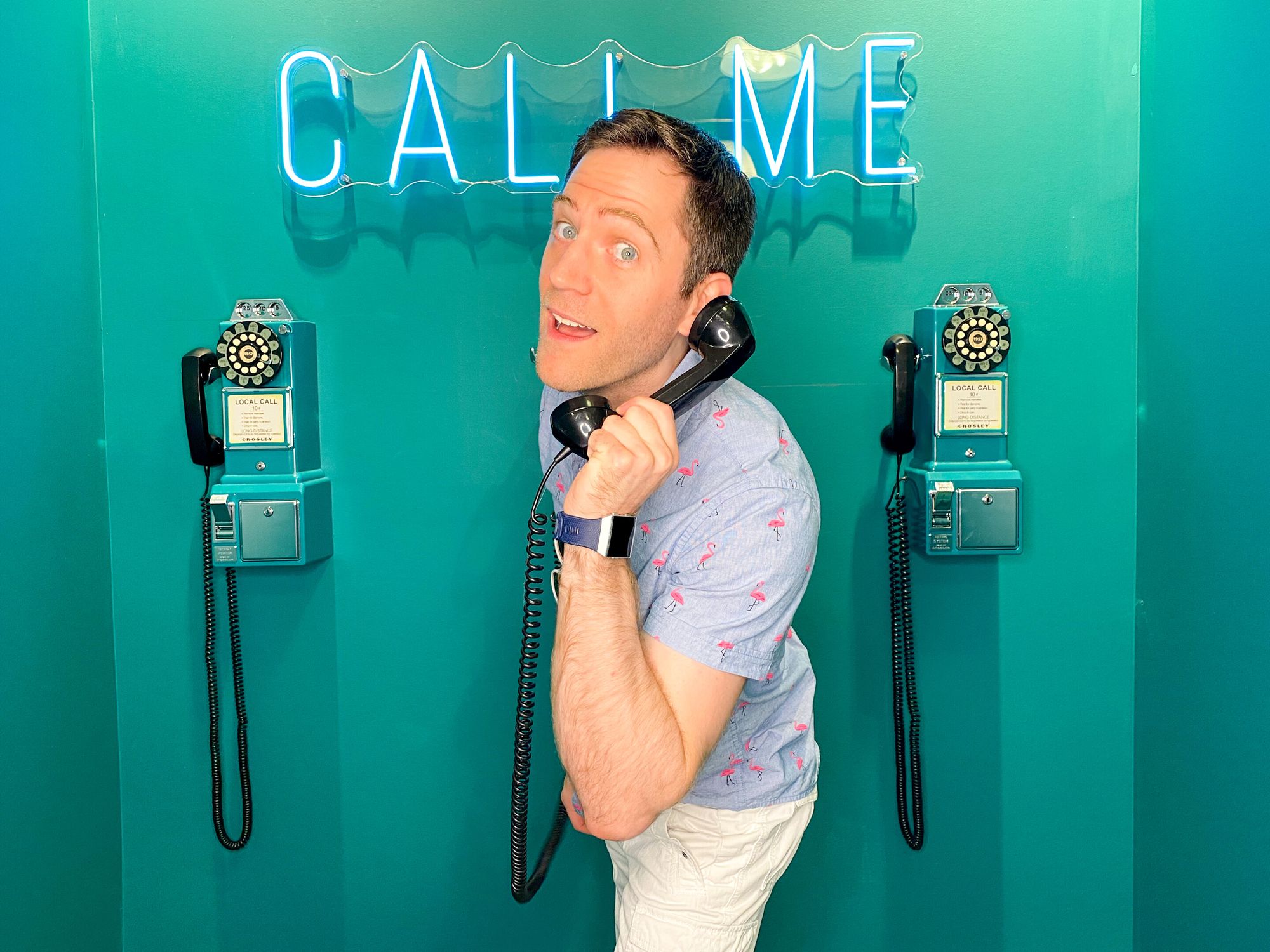 Ryan in front of three old school telephones with a receiver in his hand and leaning toward the camera.